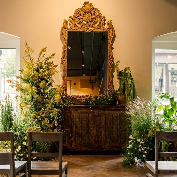 Ornate gold mirror above a wooden cabinet, flanked by lush plants and chairs, with large windows.