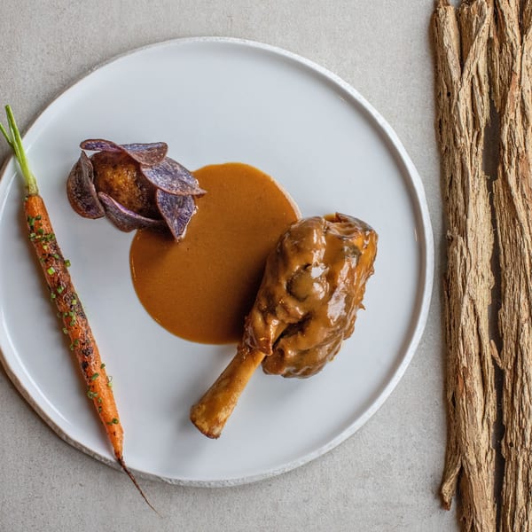 Braised lamb shank with purple potato chips, roasted carrot, and rich brown sauce on a white plate.