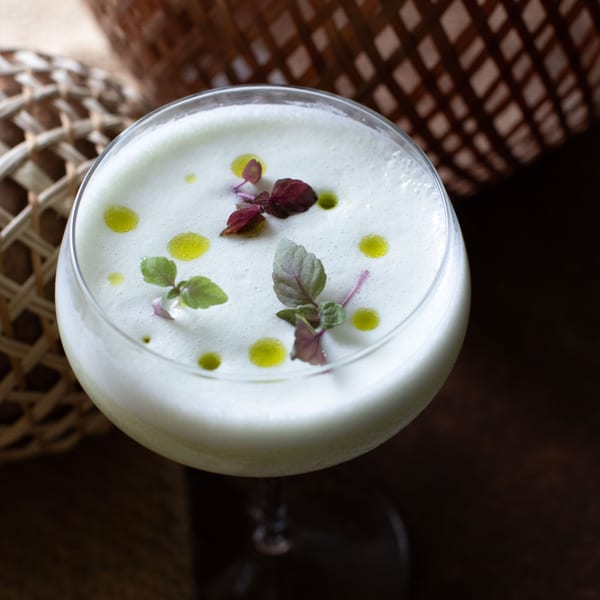 Close-up of a creamy white cocktail in a stemmed glass, garnished with green and purple herbs and oil drops.