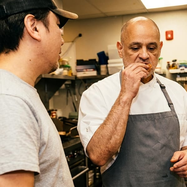 Two chefs in a commercial kitchen, one tasting food and the other observing.