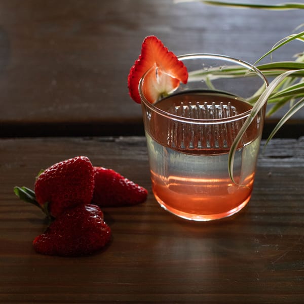 A strawberry-garnished cocktail with a pink hue sits on a dark wooden table next to fresh strawberries.