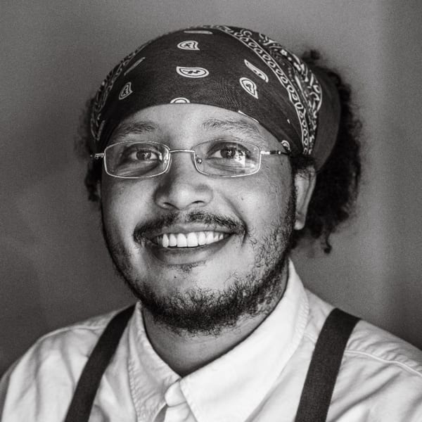 Smiling chef with a bandana, glasses, beard, white shirt, and suspenders.