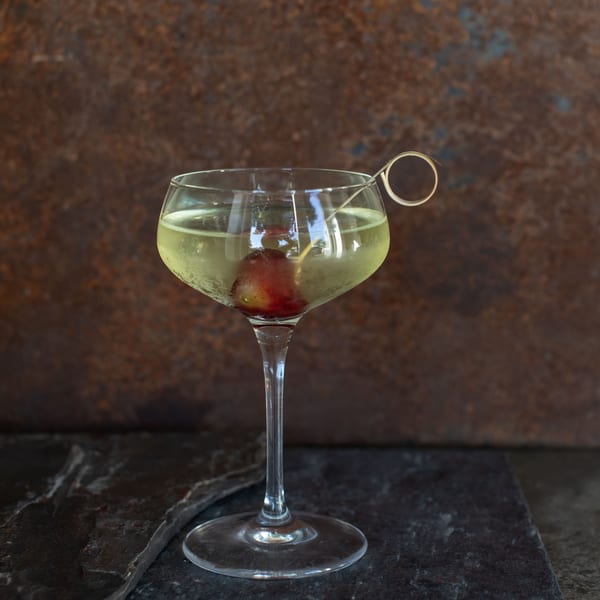 A coupe glass with a light yellow cocktail and a red grape garnish on a dark stone surface.