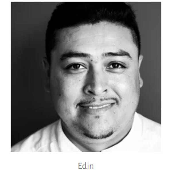 Black and white headshot of a smiling man with a mustache and goatee, wearing a chef's coat.