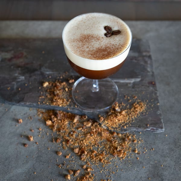 Espresso martini with foamy top, cinnamon dust, and coffee beans on a dark stone surface.
