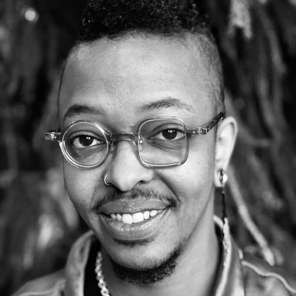 Black and white close-up of a smiling person with glasses, nose ring, and chain necklace.