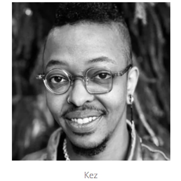Black and white portrait of Kez, a person with unique glasses and a confident smile.