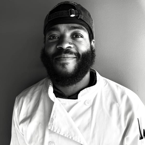 Black and white portrait of a smiling chef with a beard and baseball cap.