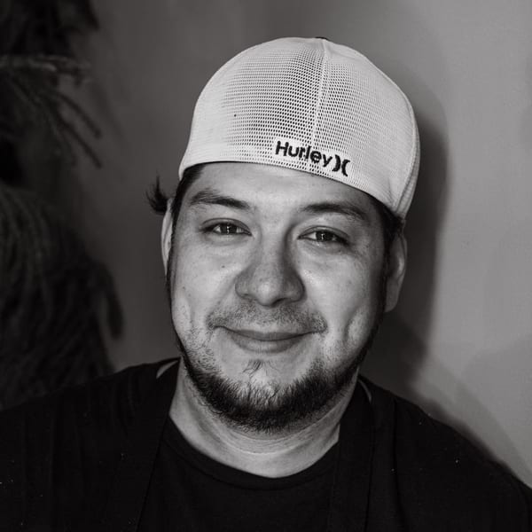 Black and white portrait of a smiling man wearing a Hurley baseball cap and a dark shirt.