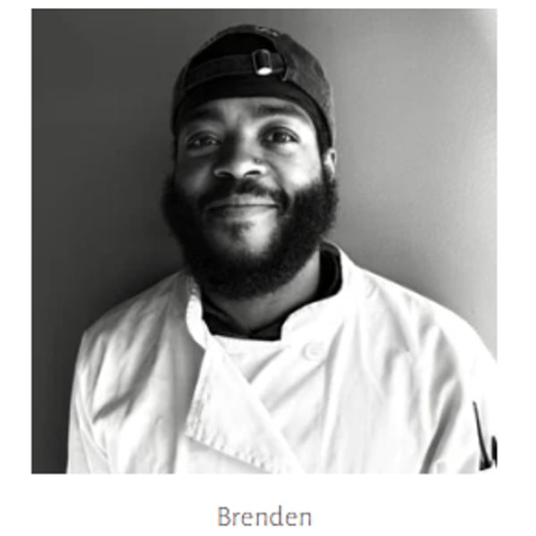 Black and white portrait of a smiling chef, Brenden, wearing a chef's coat and backward baseball cap.